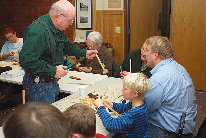 Pete Olsen (standing) gives instruction to a member of a beginning class in wood carving, at the Little Brothers, Friends of the Elderly Building.